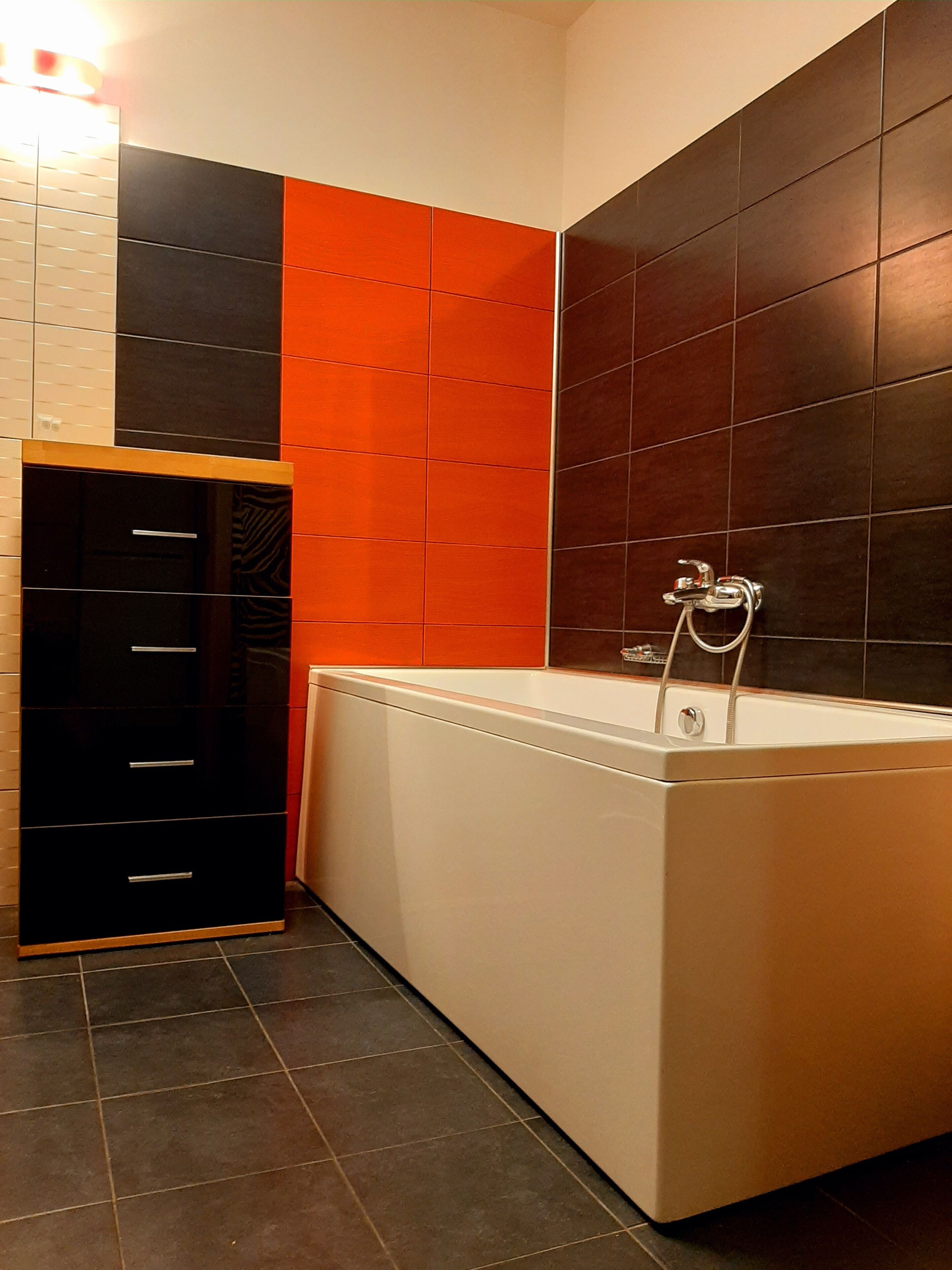 Un baño moderno con una bañera, un revestimiento de baldosas negro y naranja, y un mueble de madera.