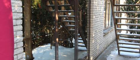 A wooden staircase leading to a glass door, a brick wall, and a wooden deck with snow on the ground.