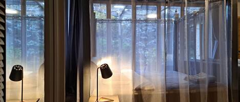 A bedroom with a bed, two chairs, a nightstand, and sheer curtains.