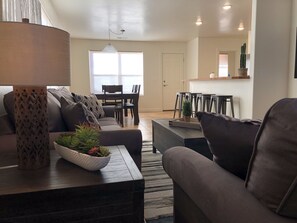 Una sala de estar moderna con un sofá, una mesa de centro y objetos decorativos. Se observa un comedor con una mesa y sillas al fondo.