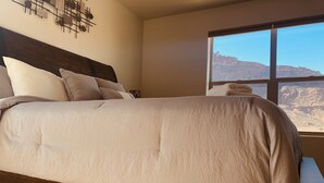 A bedroom with a wooden bed frame, a large window with a view of a mountainous landscape, and a wall-mounted abstract art piece. - Red Sand Ridge. The best place to enjoy your vacation in Moab! (Moab)