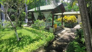 A covered outdoor seating area with a concrete path, surrounded by lush greenery and palm trees. - Explore Paradise-Palma Real cottage (Liberia)