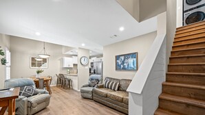 A spacious living area with a wooden staircase, a dining table, and a kitchen in the background.