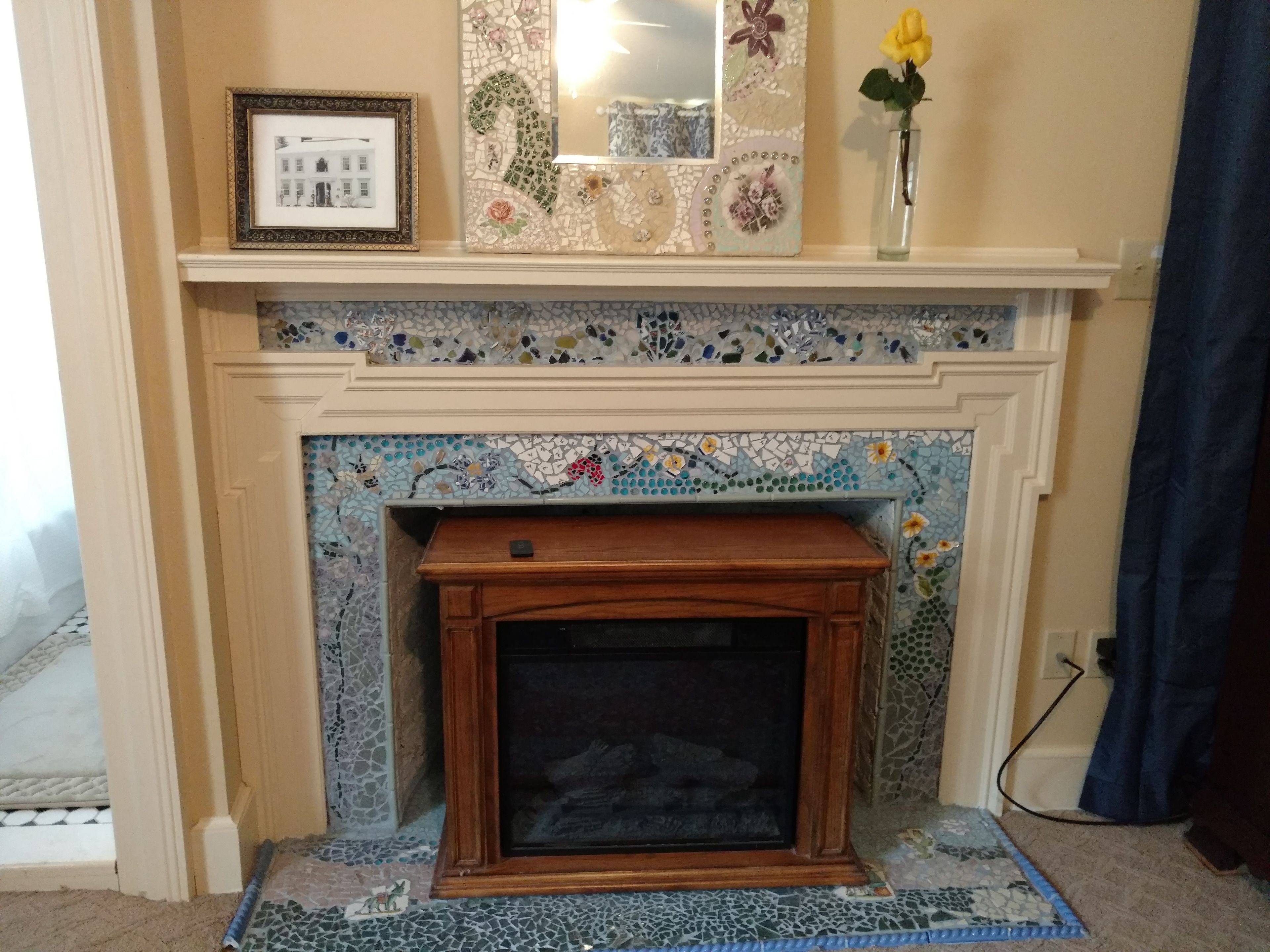 A fireplace with mosaic tile work, a framed picture, and a vase with a flower on the mantel.