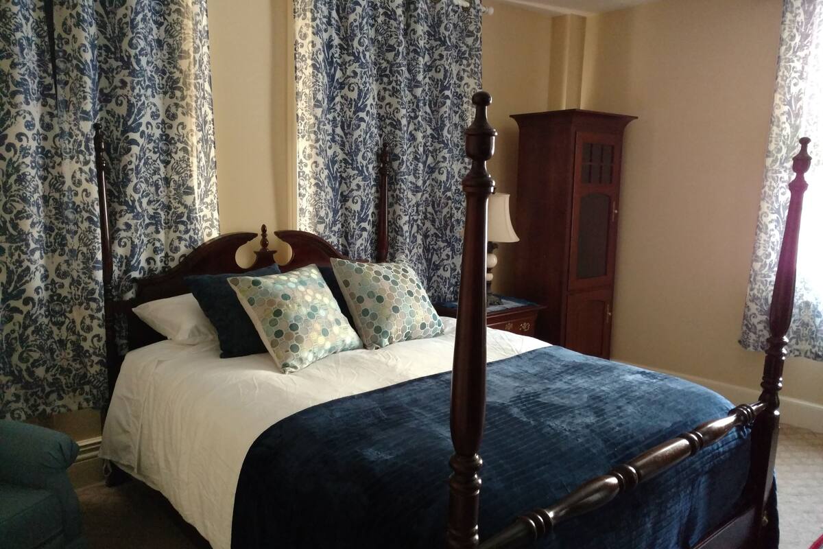 A four-poster bed with a dark wooden frame, a white and blue bedspread, and decorative pillows with a geometric pattern.