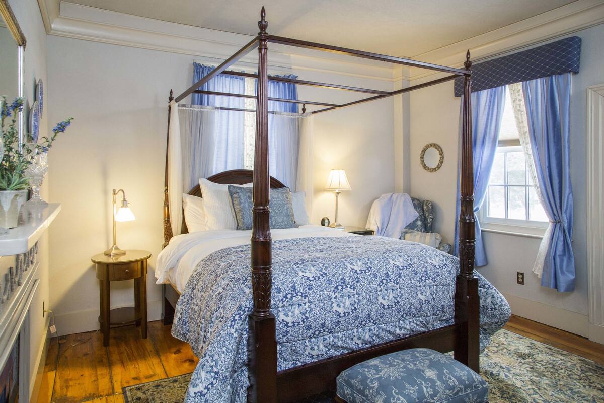 A four-poster bed with a floral bedspread, bedside tables with lamps, a mirror on the wall, and a window with curtains.