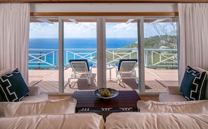 A room with a view of the ocean, featuring a sofa, two chairs, and a table with a fruit bowl.