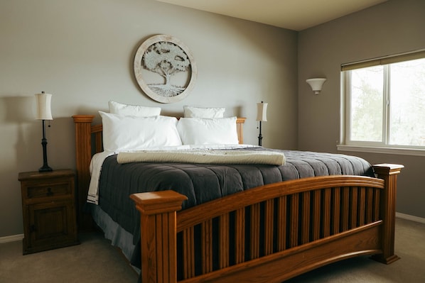 A bedroom with a wooden bed, two bedside tables, a wall-mounted lamp, and a window with a view. - Relax in beautiful mountain serenity while planning your local adventures. (Coeur d' Alene)
