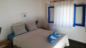 Een slaapkamer met een bed, kussens, een nachtkastje, een lamp, een raam met blauwe kaders en een gordijn.