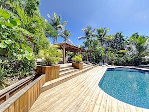 A wooden deck leading to a pool surrounded by tropical plants and a wooden pergola.