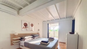 Un dormitorio con cama, mesita de noche, lámpara, aire acondicionado instalado en la pared y puerta que da a un balcón.