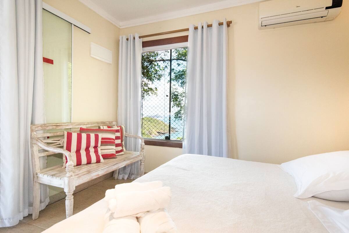 Una habitación con cama, un banco con almohadones, una ventana con cortinas y un equipo de aire acondicionado.