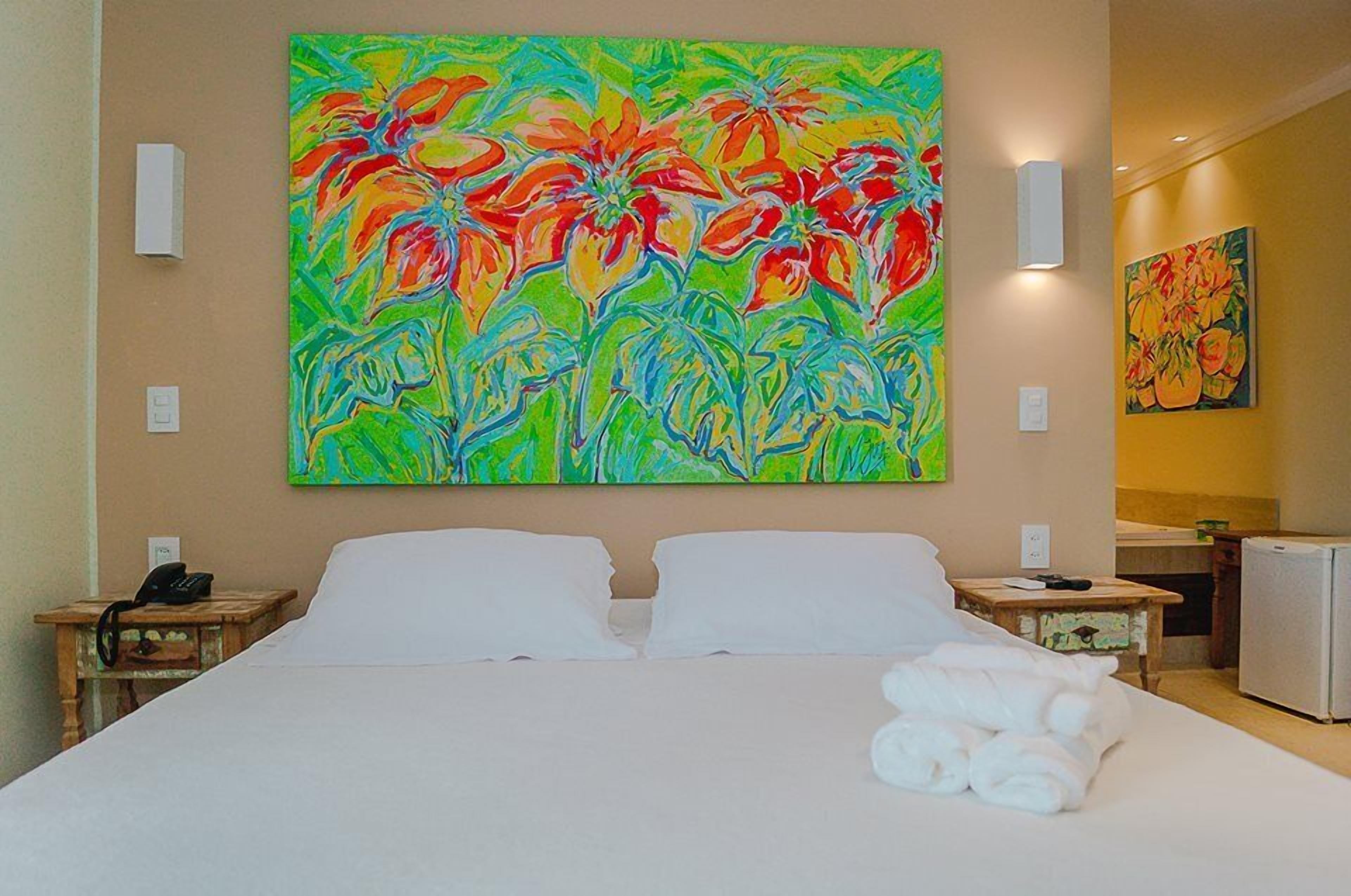 Une chambre d’hôtel avec un grand tableau coloré au mur, un lit recouvert de draps blancs et une petite table sur laquelle se trouvent un téléphone et une télécommande.