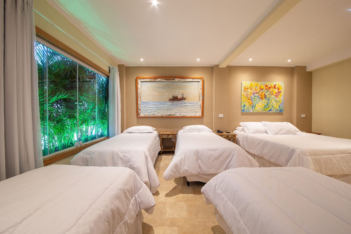 Habitación de hotel con cuatro camas individuales, un ventanal con vistas al mar, un cuadro en la pared y una mesita de noche con una lámpara.