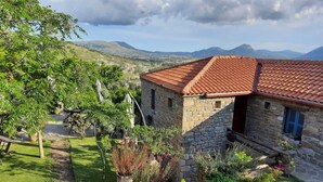 Une maison en pierre avec un toit recouvert de tuiles rouges, entourée d’un jardin bien entretenu où poussent différentes espèces de plantes, et d’où l’on peut admirer des collines lointaines.