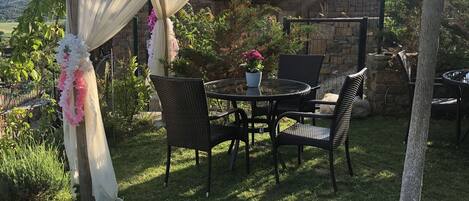 A patio area with a pergola covered in white and pink floral decorations, a round table with chairs, and a stone wall in the background.
