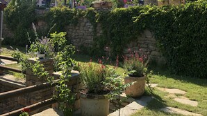 Un bâtiment en pierre, surmonté d’un toit de tuiles rouges, entouré d’une végétation luxuriante et agrémenté de plantes en pot.