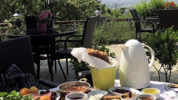 Une table dressée avec divers plats et boissons, notamment des olives, des tomates et du pain, dans un cadre extérieur agrémenté de chaises et offrant une vue sur la verdure.