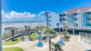 Un hotel frente a la playa con varios balcones, un área de piscina con sombrillas y un paseo marítimo que conduce a la playa.