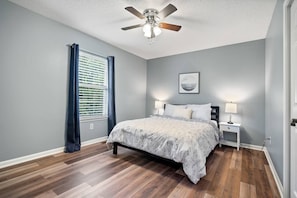 Ein Schlafzimmer mit einem Bett, einem Nachttisch, einem Deckenventilator, einem Fenster mit Vorhängen und einem Wandbild.