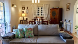 A living room with a sofa, a dining table, and a cabinet. - Casa das Rosas, a peaceful stress-free villa to relax (Santa Bárbara de Nexe)