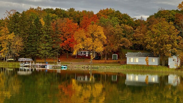 A lakeside cabin with a dock and boats, surrounded by autumn foliage.