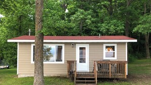A small, single-story cabin with a red roof, a front door, and a small porch with steps leading up to it.