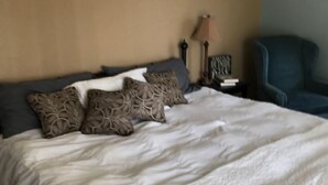 A neatly made bed with patterned pillows, a bedside table with a lamp, and a chair in a room with beige walls.