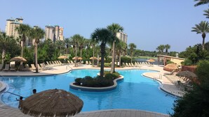 Área de piscina en un resort con múltiples sillas de descanso y palmeras, rodeada de edificios altos.