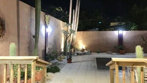 Un patio exterior nocturno con cactus y plantas en macetas, bancos de madera y una pared decorada con una calavera de cactus.