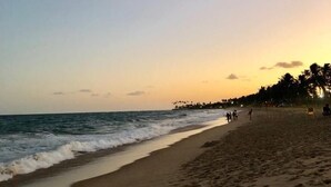Une plage où l’on voit des empreintes de pas menant à l’eau, une côte lointaine bordée de palmiers et un ciel dégagé.