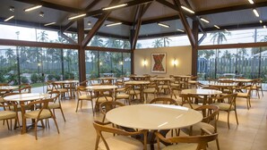Un vaste espace de restauration intérieur, doté de plusieurs tables rondes et de chaises en bois, de grandes fenêtres offrant une vue sur les palmiers et d’un haut plafond avec des poutres apparentes.