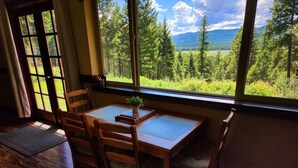 Huoneessa on puinen pöytä ja tuolit, ikkunasta avautuu näkymä metsään ja vuorille, ja ovessa on verhot.
