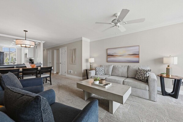 A modern living room with a sofa, coffee table, and chairs. There is a dining area with a table and chairs. A ceiling fan is visible.