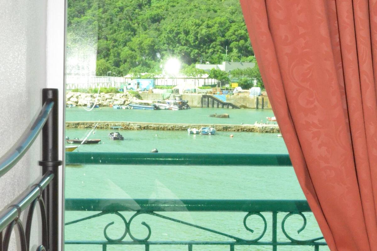 Un balcón con vista a un puerto, barandilla verde y una mesa con un vaso.