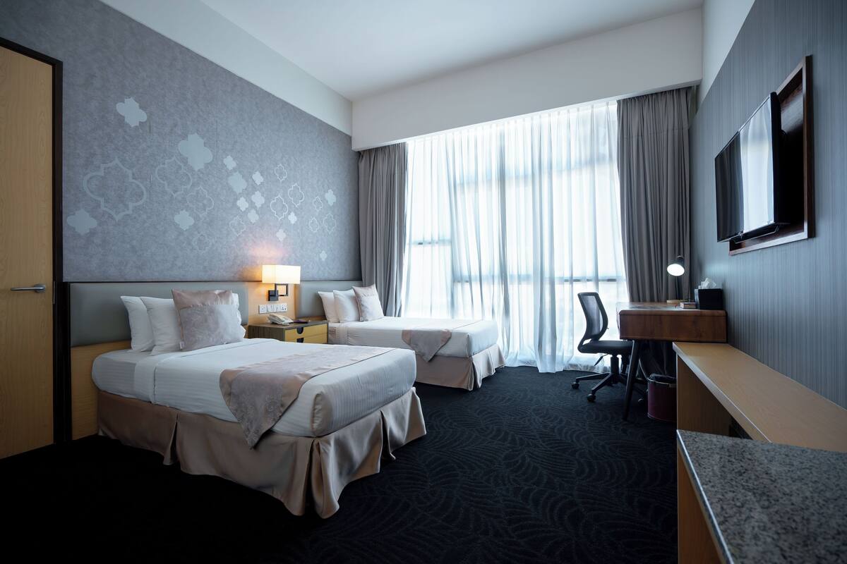 A hotel room with two beds, a desk, a chair, a TV, and a large window with curtains.