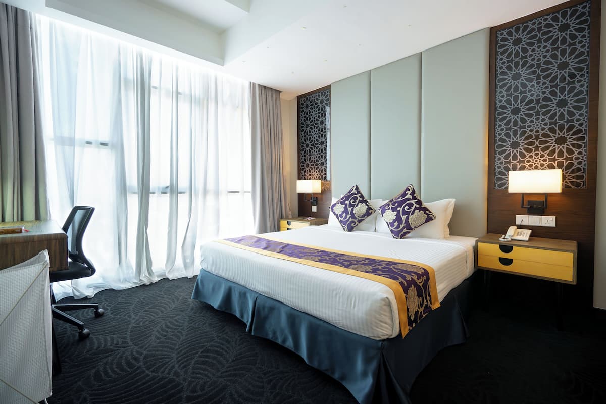 A hotel room with a large bed, a desk, a chair, and a nightstand with a lamp.