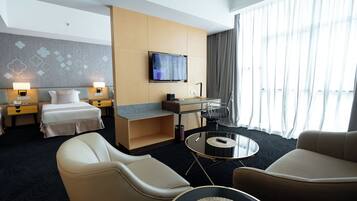 Quarto de hotel com uma cama, duas mesas de cabeceira com luminárias, uma TV, uma escrivaninha com cadeira, uma mesa de centro redonda e duas poltronas.