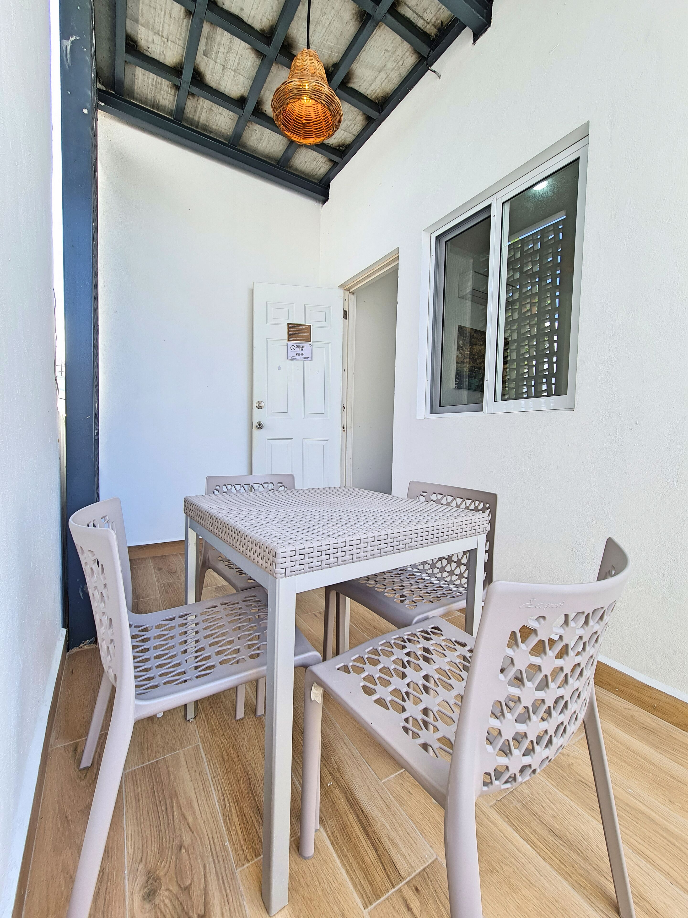 A room with a white door, a window with a metal grill, a woven light fixture, and a table with perforated chairs.