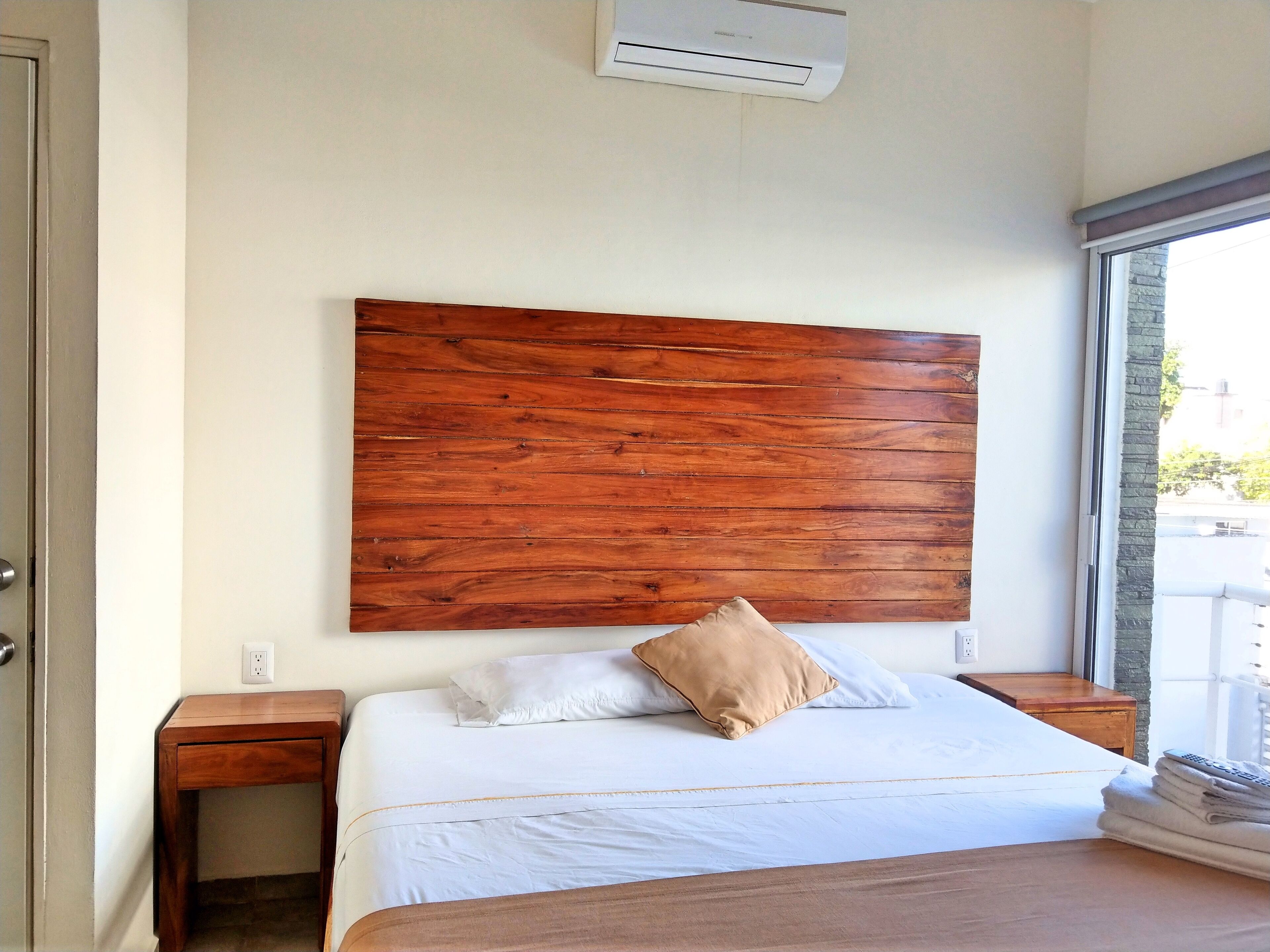 A bedroom with a wooden headboard, a white bedspread, a beige pillow, two bedside tables, and a window with a view.