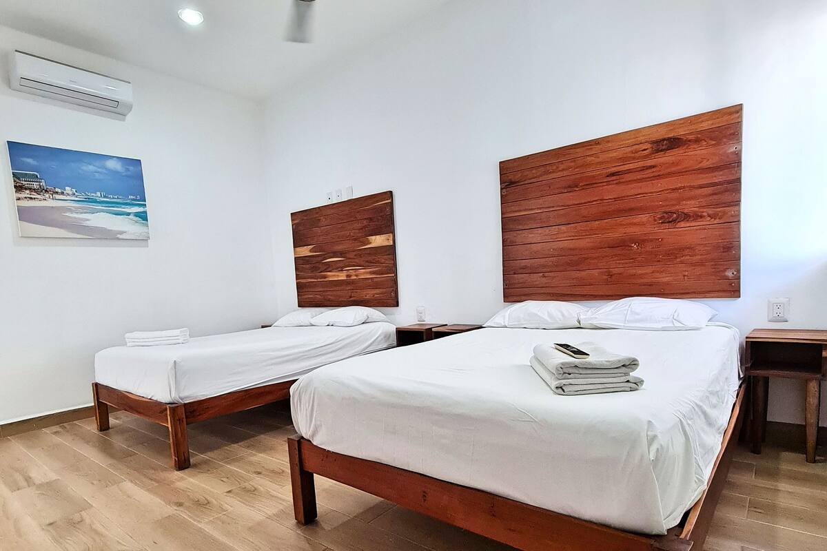 Une chambre d’hôtel avec deux lits en bois, du linge de lit blanc, un ventilateur de plafond, un appareil de climatisation et un tableau sur le thème de la plage accroché au mur.