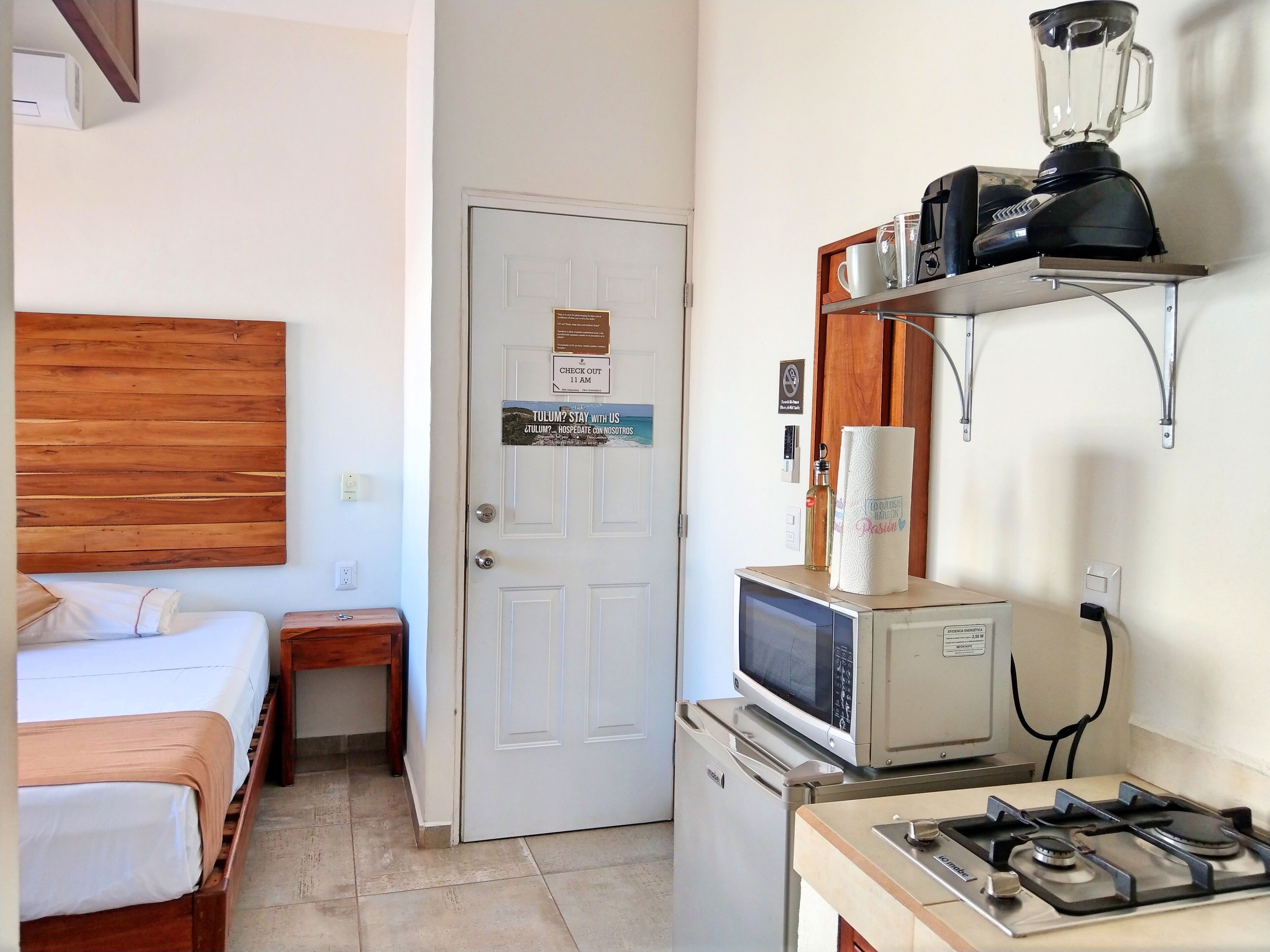 A compact hotel room with a bed, a small bedside table, a microwave, a gas stove, and a shelf with a blender.