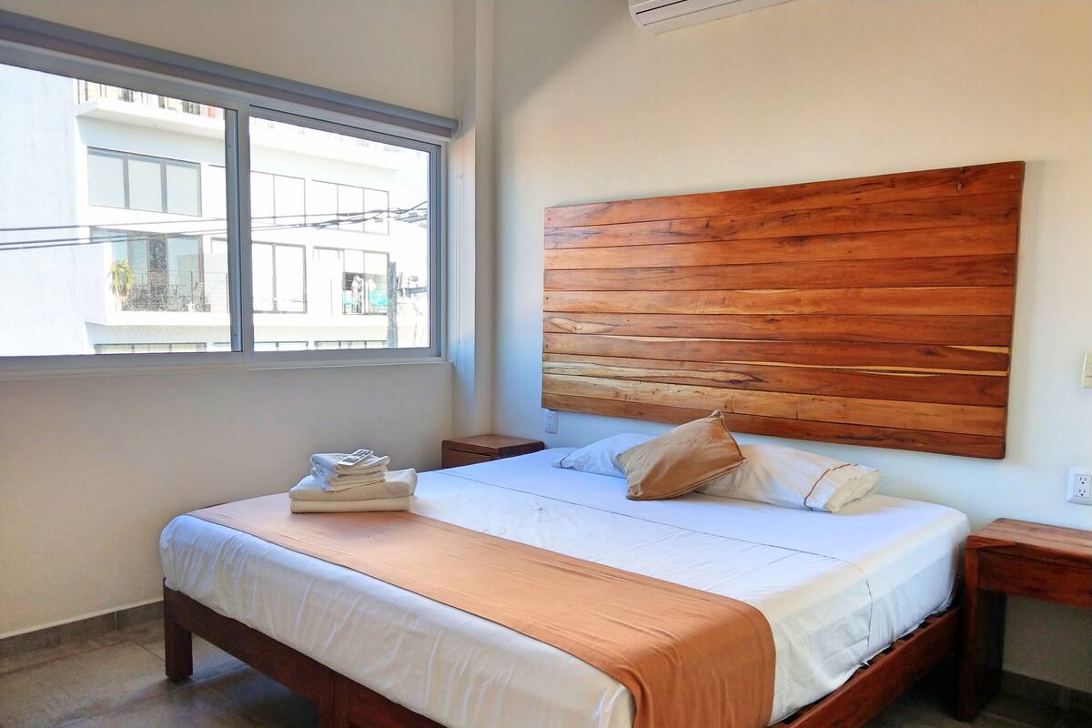 Une chambre avec un grand lit, une tête de lit en bois, une fenêtre et une vue sur les bâtiments environnants.