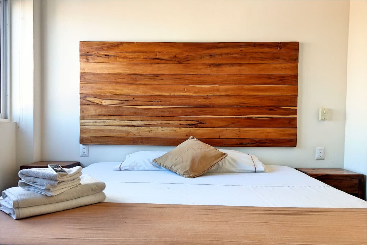 Un lit bien fait, avec une tête de lit en bois, une table de chevet sur laquelle se trouvent une télécommande et des serviettes, et une prise électrique au mur.