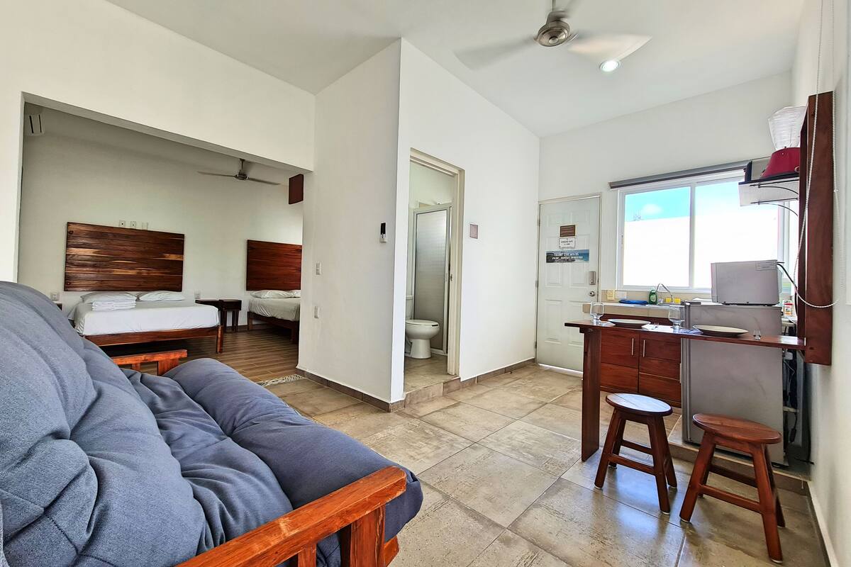 Un vaste espace de vie comprenant un grand canapé, un lit avec une tête de lit en bois, une kitchenette équipée d’un réfrigérateur et une salle de bain avec des toilettes.