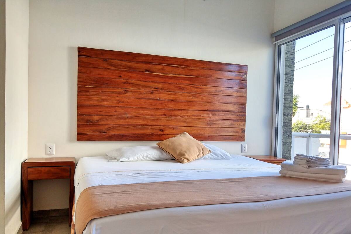 Une chambre à coucher avec une tête de lit en bois, un lit recouvert de draps blancs, un oreiller beige et une fenêtre donnant sur des immeubles.