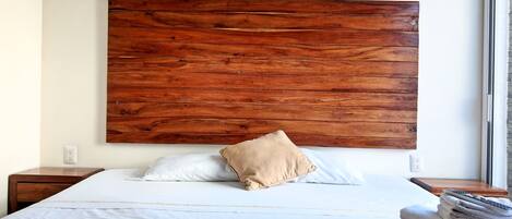Una cama bien tendida, con un cabecero de madera, un cojín color beige y una pila de toallas a un lado.