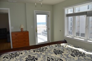 Een slaapkamer met een bed, een dressoir, een deur die leidt naar een balkon en een raam met uitzicht op het strand.