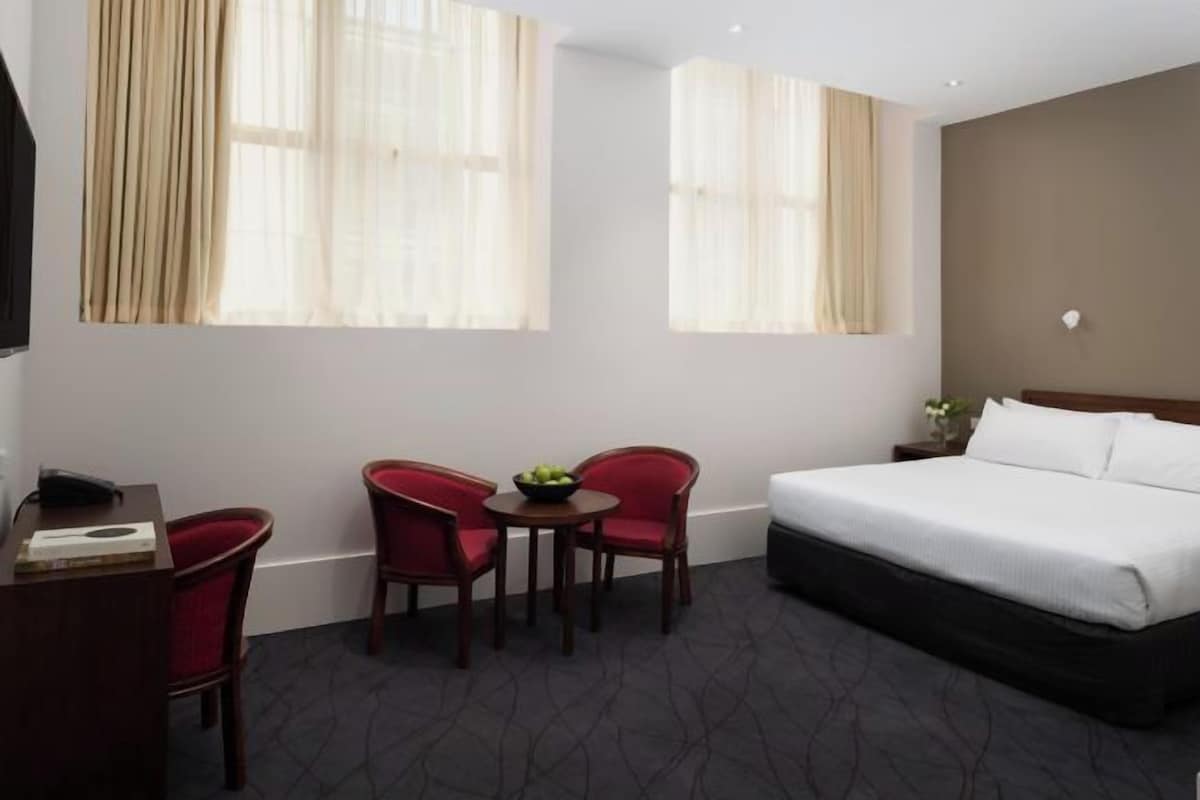 Habitación de hotel con una cama, dos sillas rojas, una mesita con un frutero y un escritorio con un teléfono.