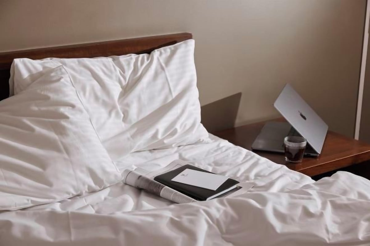 Una cama bien hecha con sábanas blancas, una computadora portátil sobre una mesita de noche de madera y un vaso de agua.